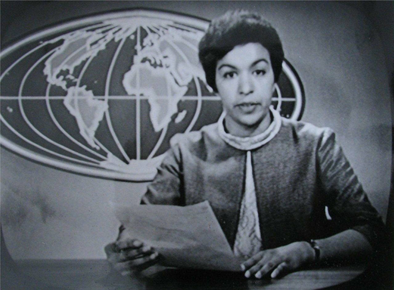 Black-and-white still of Eugènie reading the news in front of the world map of the NTS news, 1965